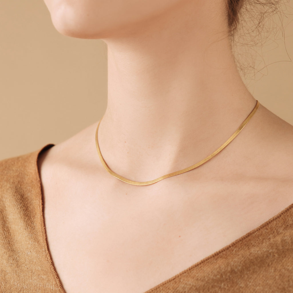 Delicate gold herringbone chain necklace resting flat along the collarbone, worn with a neutral toned V neck top against a beige background.