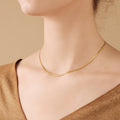 Delicate gold herringbone chain necklace resting flat along the collarbone, worn with a neutral toned V neck top against a beige background.