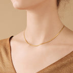 Delicate gold herringbone chain necklace resting flat along the collarbone, worn with a neutral toned V neck top against a beige background.