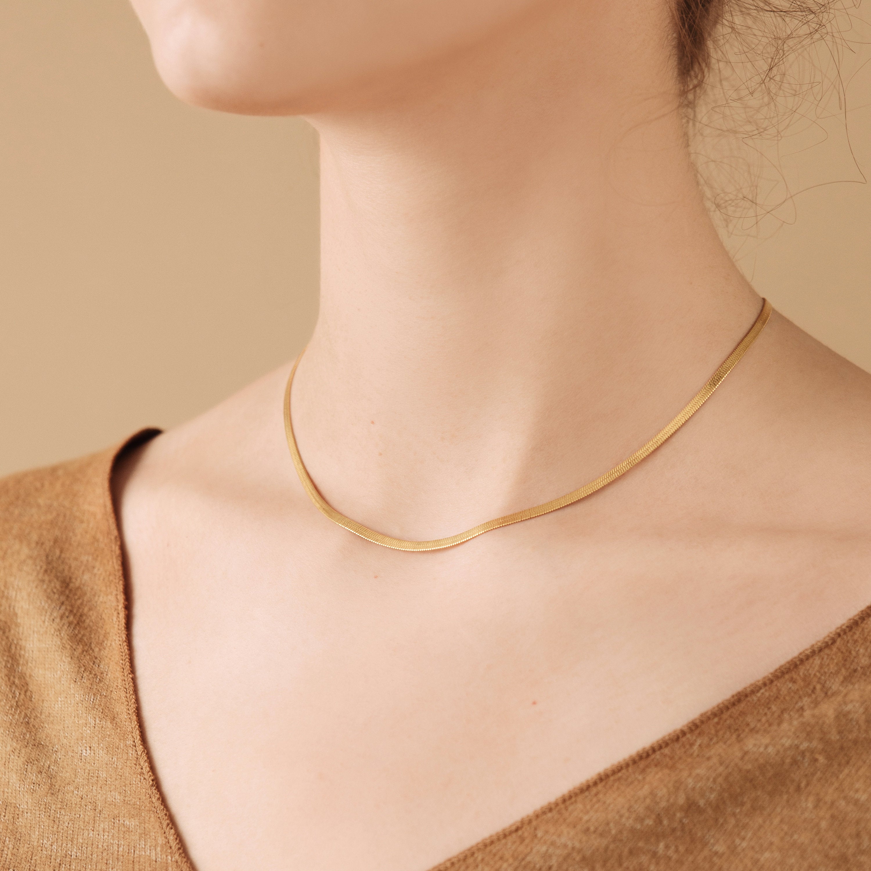 Delicate gold herringbone chain necklace resting flat along the collarbone, worn with a neutral toned V neck top against a beige background.