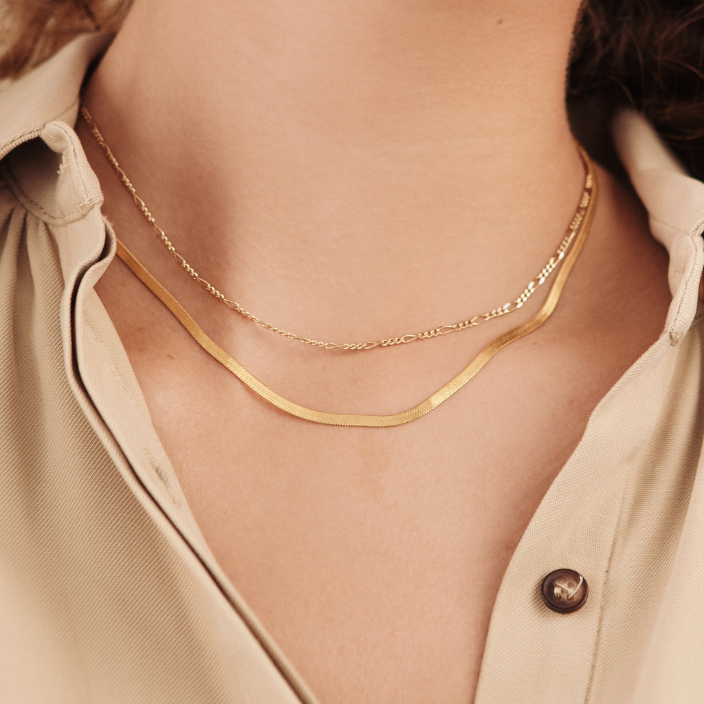A model wears a layered necklace look featuring a flat, gold herringbone chain and a delicate Figaro chain