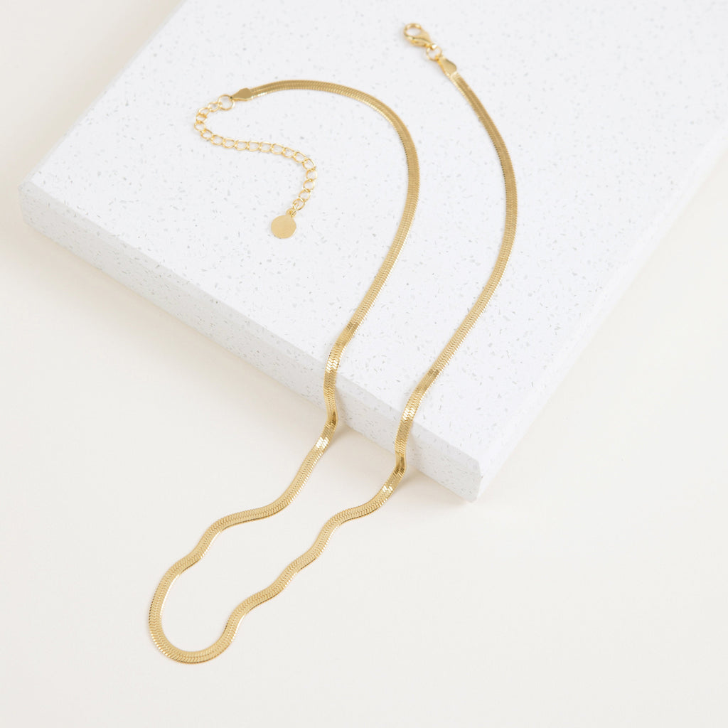 A close-up view of a gold herringbone necklace resting on a terrazzo display block.