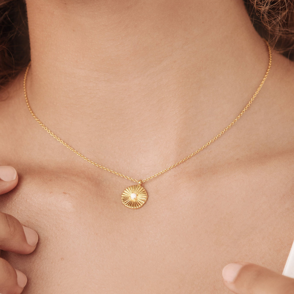 The Nova Necklace, a gold sunburst pendant with vintage-inspired design, rests on a woman's neck as her hands gently touch the chain.