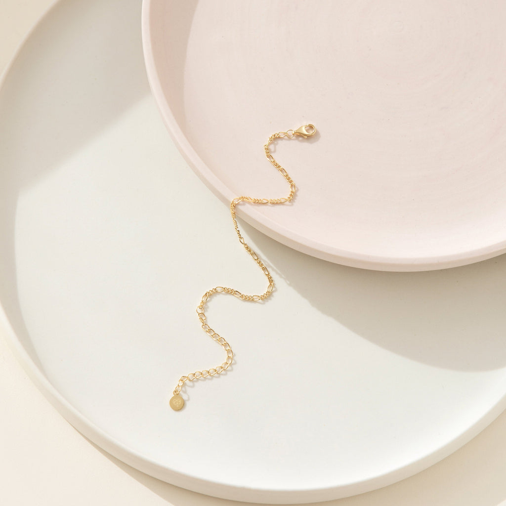The Figaro Bracelet features a gold unisex chain design, artfully displayed on stacked round white and pale pink ceramic plates—modern jewelry elegance in every detail.