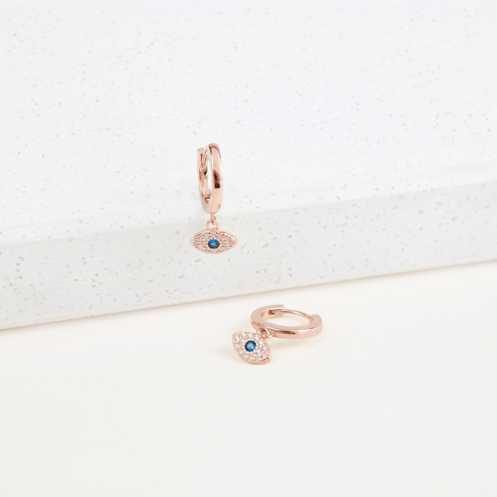 Iris Huggies—rose gold earrings with blue gemstone evil eye charms—bring a modern, stylish twist to protection jewelry, shown atop a white speckled surface.