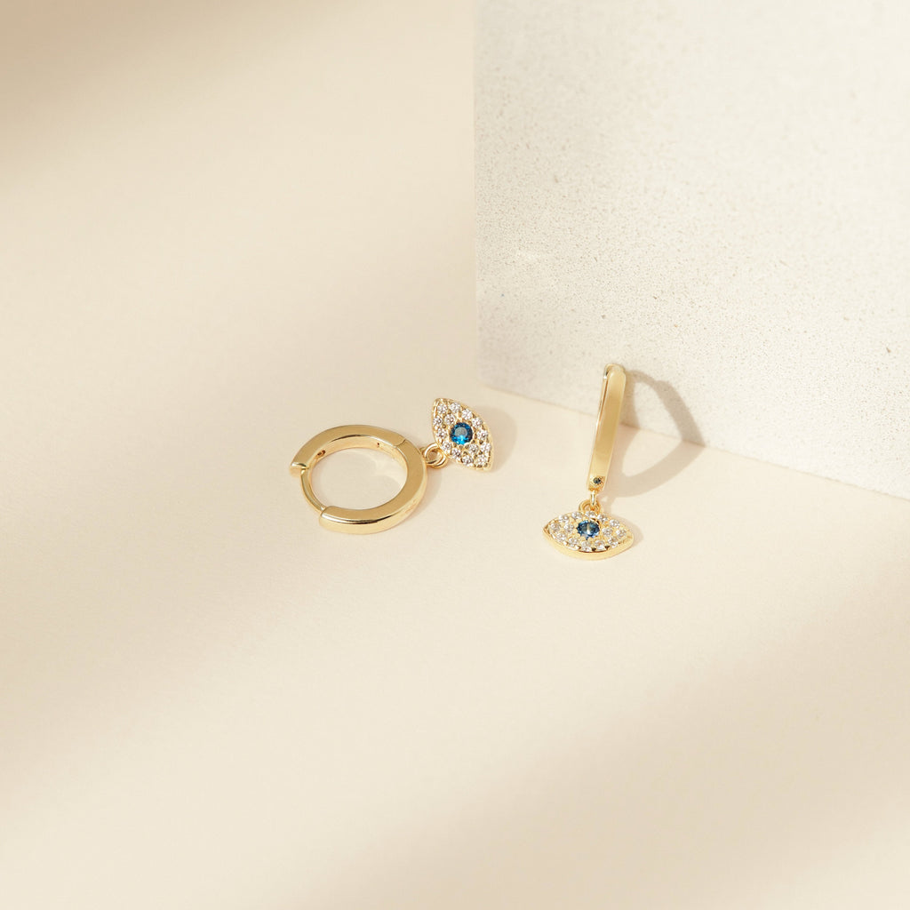Iris Huggies are gold earrings featuring blue and white evil eye charms, symbolizing protection jewelry and shown on a light beige background.