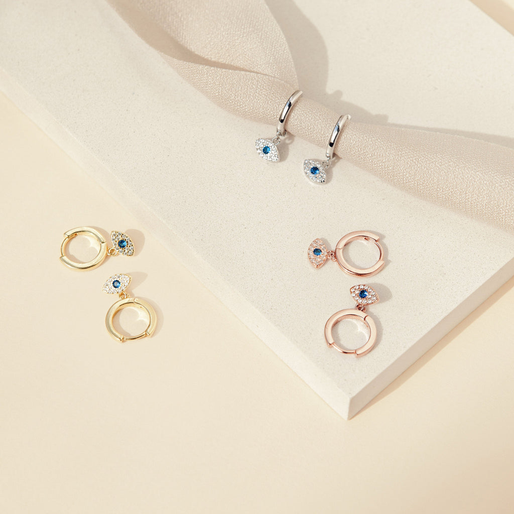 The Iris Huggies: three pairs of silver, gold, and rose gold huggie earrings with blue stone charms on a beige background—ideal for fans of stylish protection jewelry.