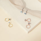 The Iris Huggies: three pairs of silver, gold, and rose gold huggie earrings with blue stone charms on a beige background—ideal for fans of stylish protection jewelry.