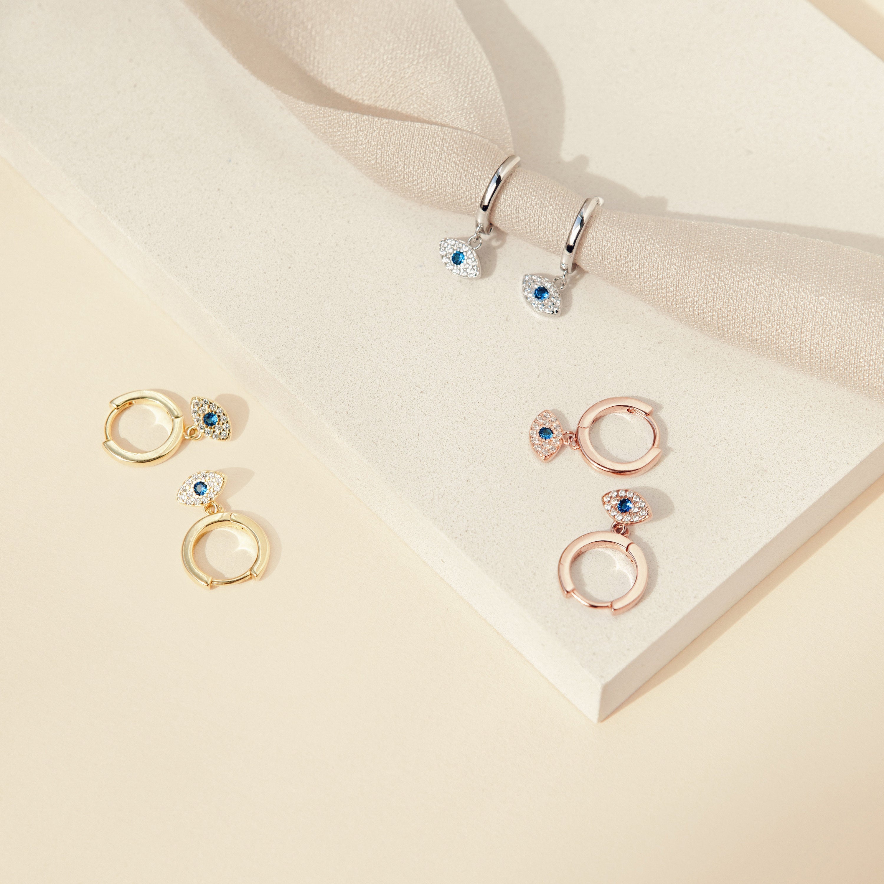 The Iris Huggies: three pairs of silver, gold, and rose gold huggie earrings with blue stone charms on a beige background—ideal for fans of stylish protection jewelry.
