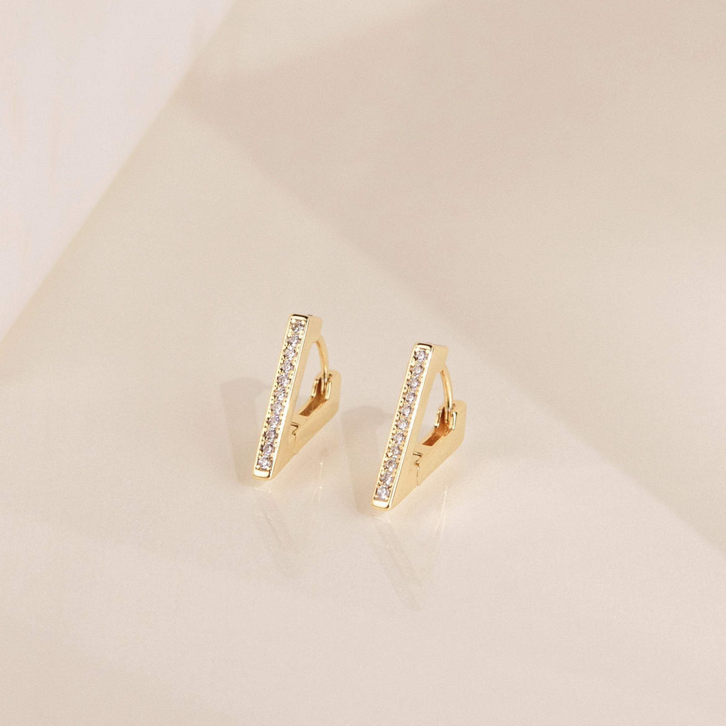 Ezra Triangle Earrings: gold geometric earrings with a vertical bar design and a pave of sparkling crystals, displayed on a beige surface.