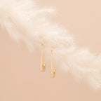 Pave Safety Pin Earrings with small crystals hang on a soft, white feather against a beige background.