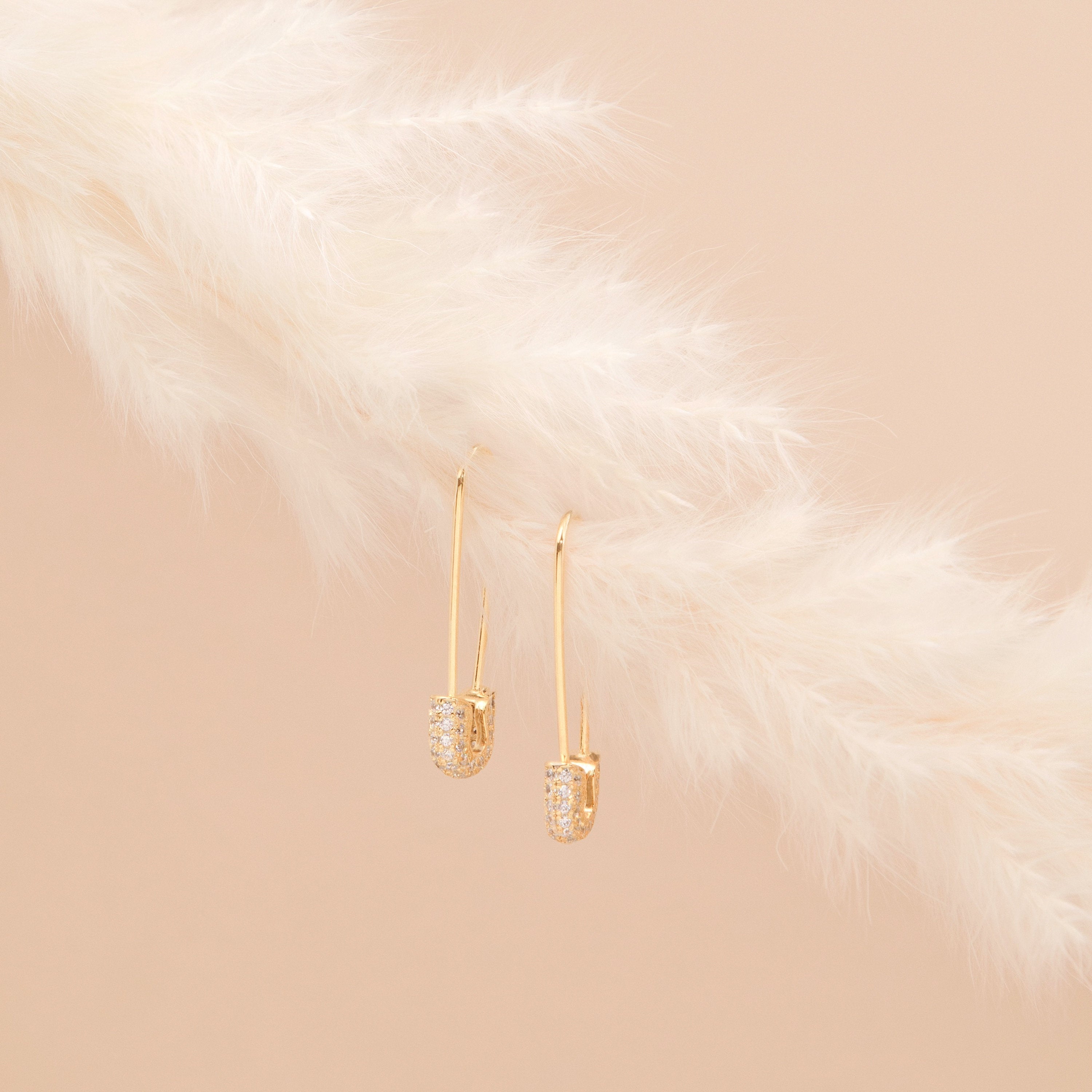 Pave Safety Pin Earrings with small crystals hang on a soft, white feather against a beige background.