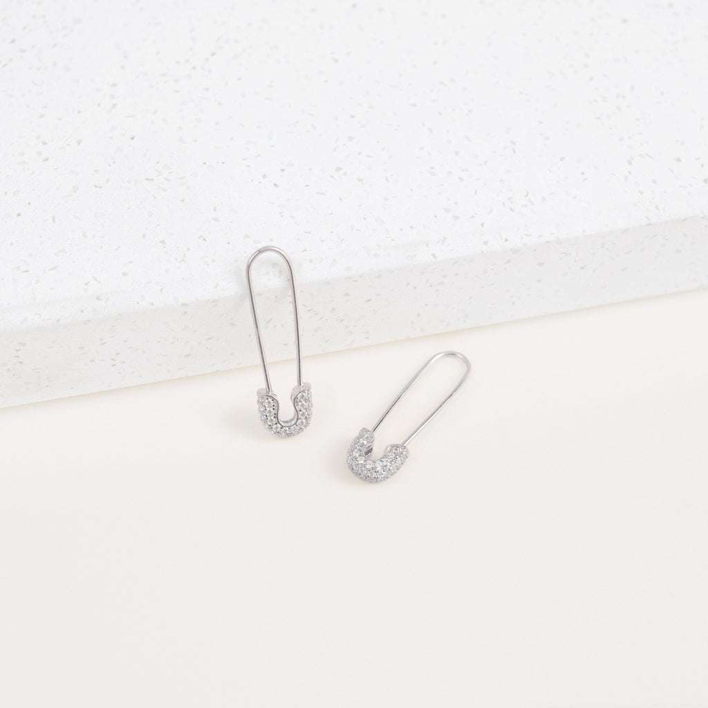 Two Pave Safety Pin Earrings with sparkling crystal accents rest on a white textured surface, adding a unique and eye-catching touch to any outfit.