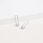 Two Pave Safety Pin Earrings with sparkling crystal accents rest on a white textured surface, adding a unique and eye-catching touch to any outfit.