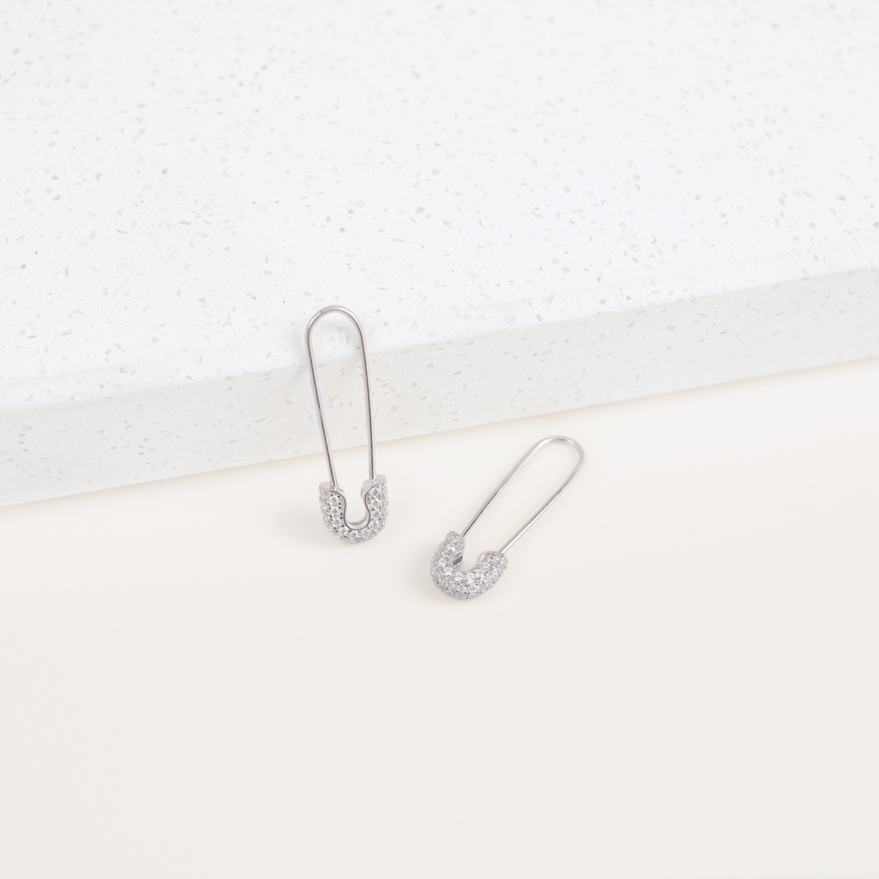 Two Pave Safety Pin Earrings with sparkling crystal accents rest on a white textured surface, adding a unique and eye-catching touch to any outfit.