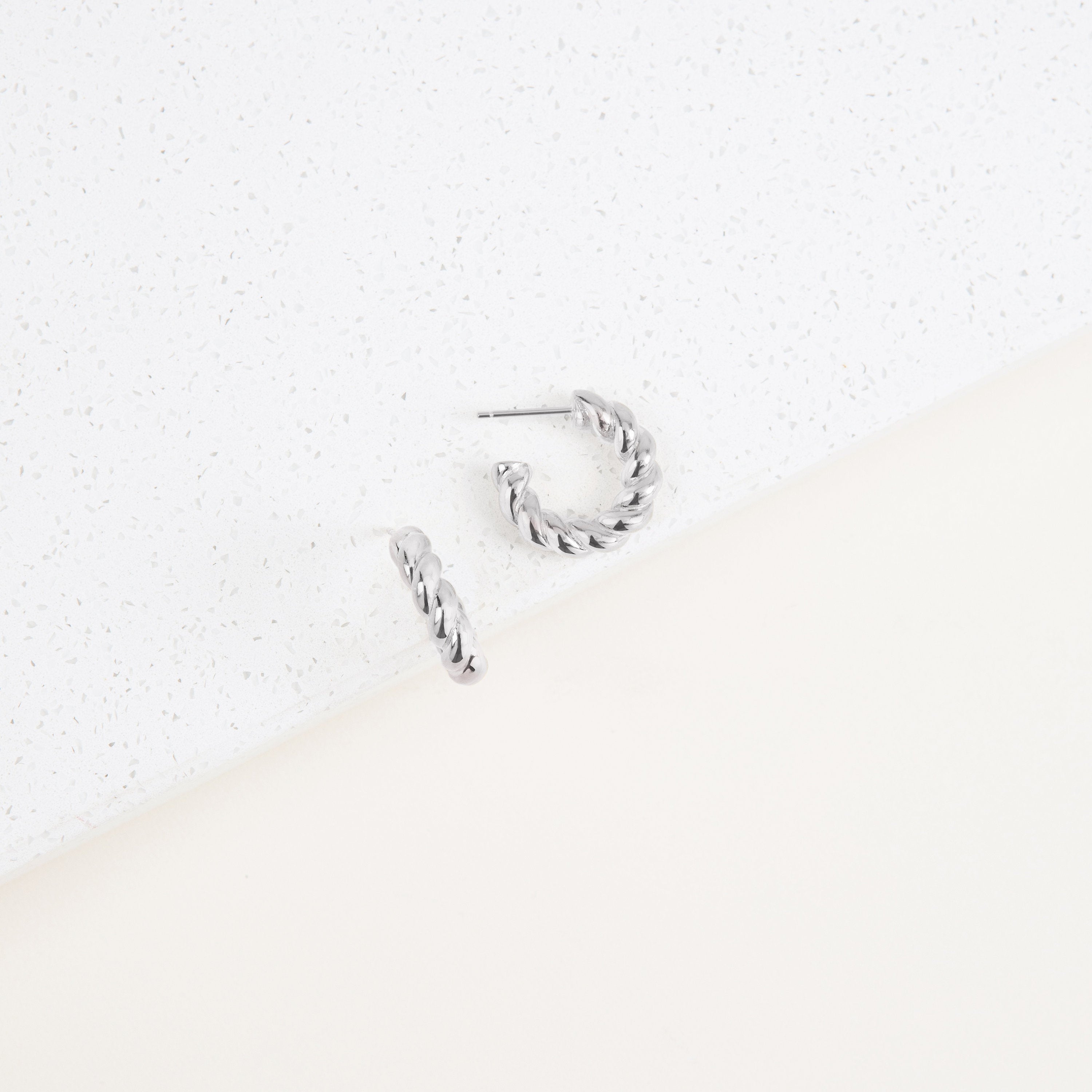 The Remi Twist Hoops are small, twisted silver hoops displayed on a white textured surface—perfect minimal jewelry for everyday wear.