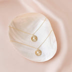 The Nova Necklace, a gold opal medallion with a sunburst design, rests on a shiny shell dish atop pink satin—perfect vintage-inspired jewelry to elevate any collection.