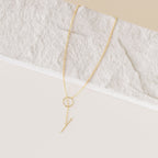 The Skyy Toggle Necklace is a minimal gold piece with a circular loop and vertical bar pendant, elegantly displayed on a lightly textured surface.