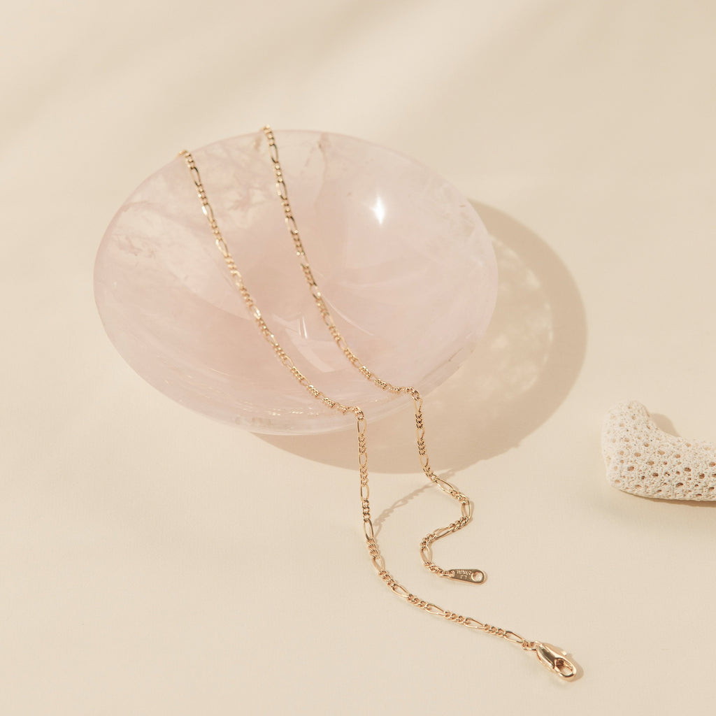 A gold Figaro chain necklace elegantly draped over a polished, rose quartz bowl. The alternating pattern of short and elongated oval links is distinct, adding visual texture and a classic Italian-inspired design to the fine gold piece.