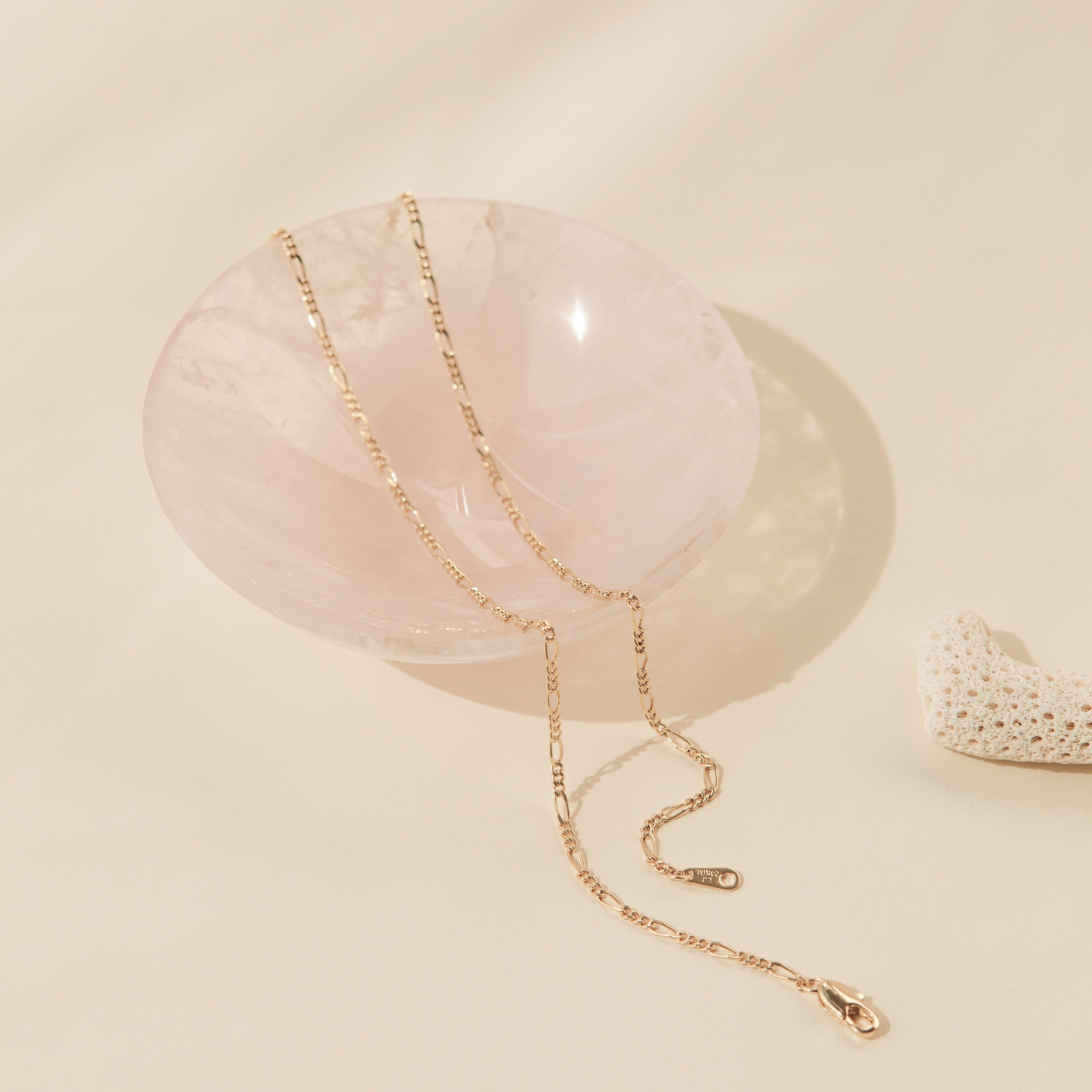 A gold Figaro chain necklace elegantly draped over a polished, rose quartz bowl. The alternating pattern of short and elongated oval links is distinct, adding visual texture and a classic Italian-inspired design to the fine gold piece.
