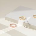 A flat lay of Dome Rings in gold, silver, and rose gold finishes arranged on a white platform, highlighting their curved, glossy surfaces.