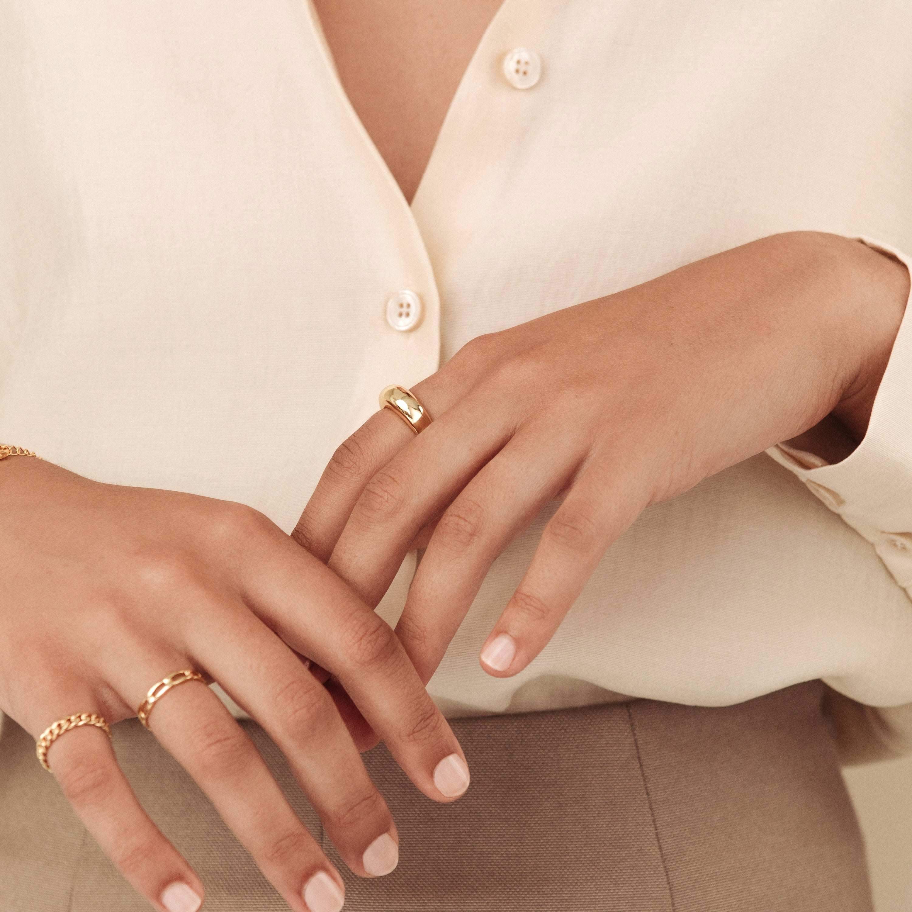 Close-up of a woman's hands adorned with the gold Dome Ring and coordinating gold bands, styled with a cream blouse and taupe trousers for an elegant, layered look.
