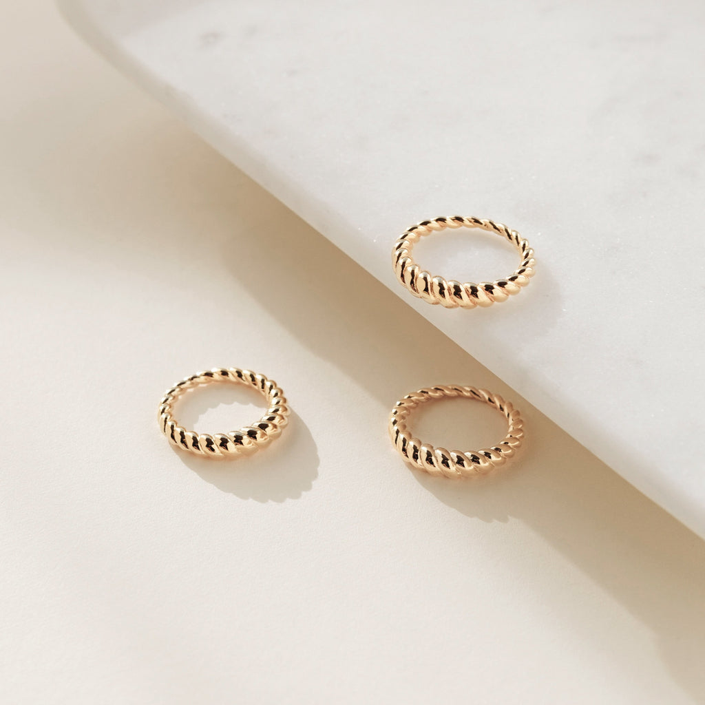 The Twisted Dome Ring, a textured gold accessory, is showcased alongside two twisted gold rings on a light marble surface with a cream background.