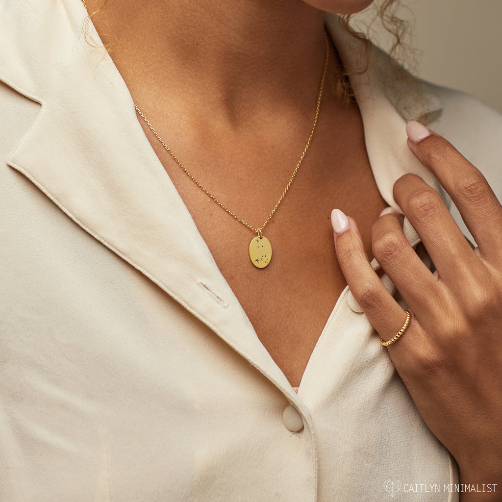 A woman in a cream blouse wears the Zodiac Constellation Necklace, featuring a round pendant, along with a matching gold ring.