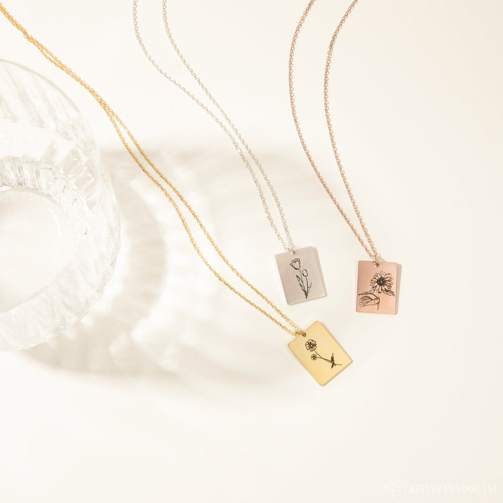 Three Birth Flower Tag Necklaces—gold, silver, and rose gold—with delicate floral engravings rest on a white surface. Perfect personalized birth month jewelry.