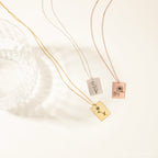 Three Birth Flower Tag Necklaces—gold, silver, and rose gold—with delicate floral engravings rest on a white surface. Perfect personalized birth month jewelry.