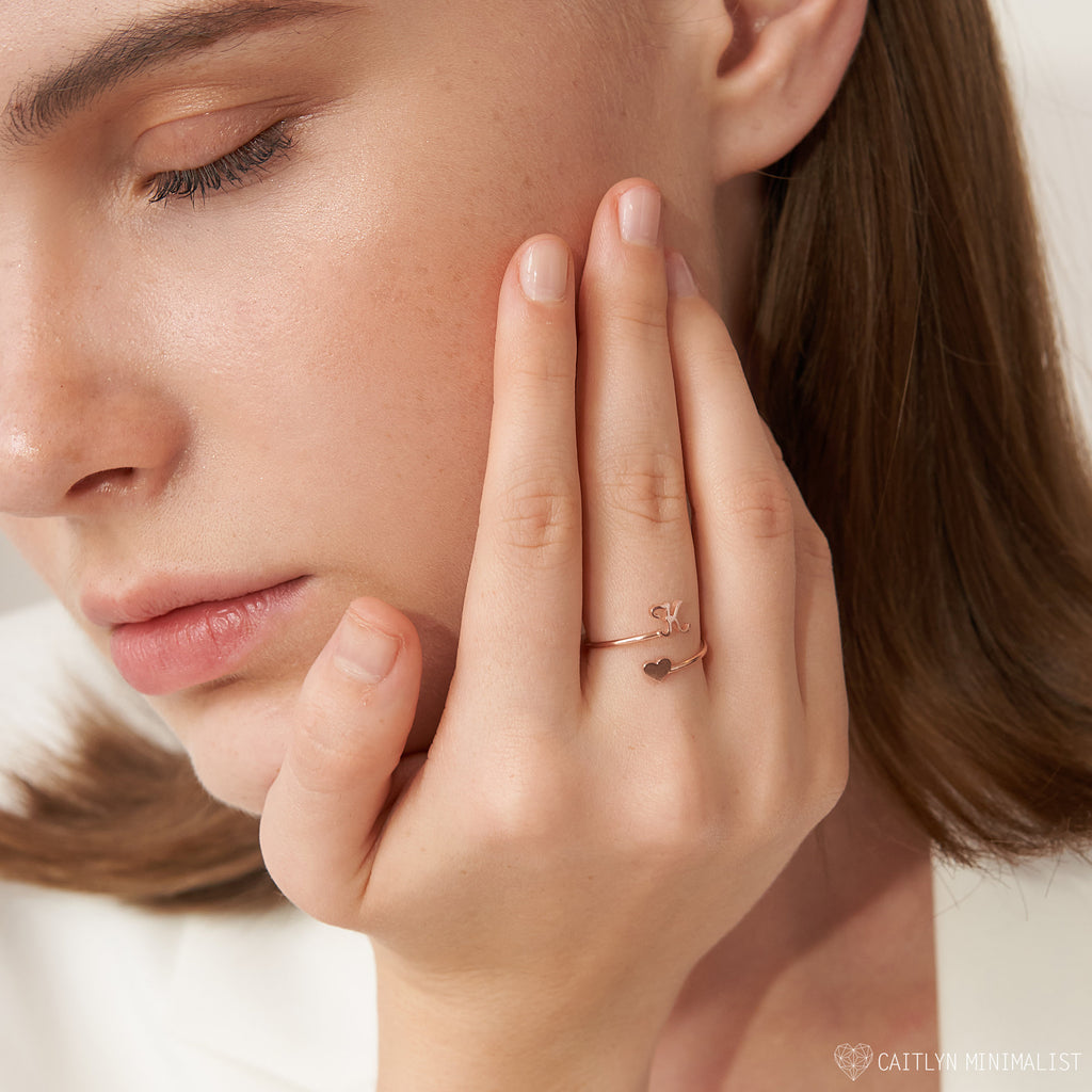 A woman touches her face, wearing the Initial Heart Wrap Ring—this customizable rose gold ring features initials, creating a unique and personal symbol of love on her finger.