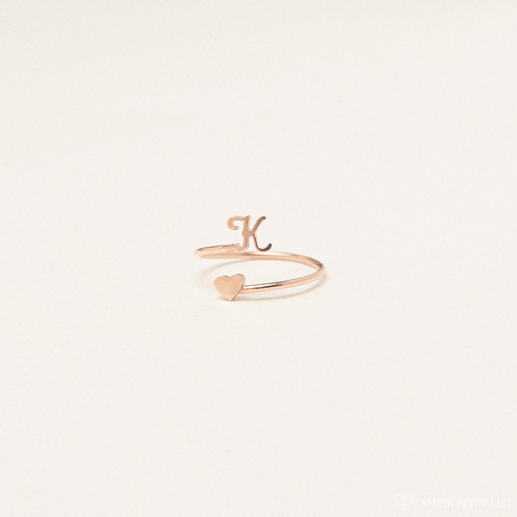 The Initial Heart Wrap Ring features a gold band with a letter K and a heart on opposite ends—an elegant symbol of love. Personalize it with any initial for a unique touch, beautifully displayed against a plain backdrop.