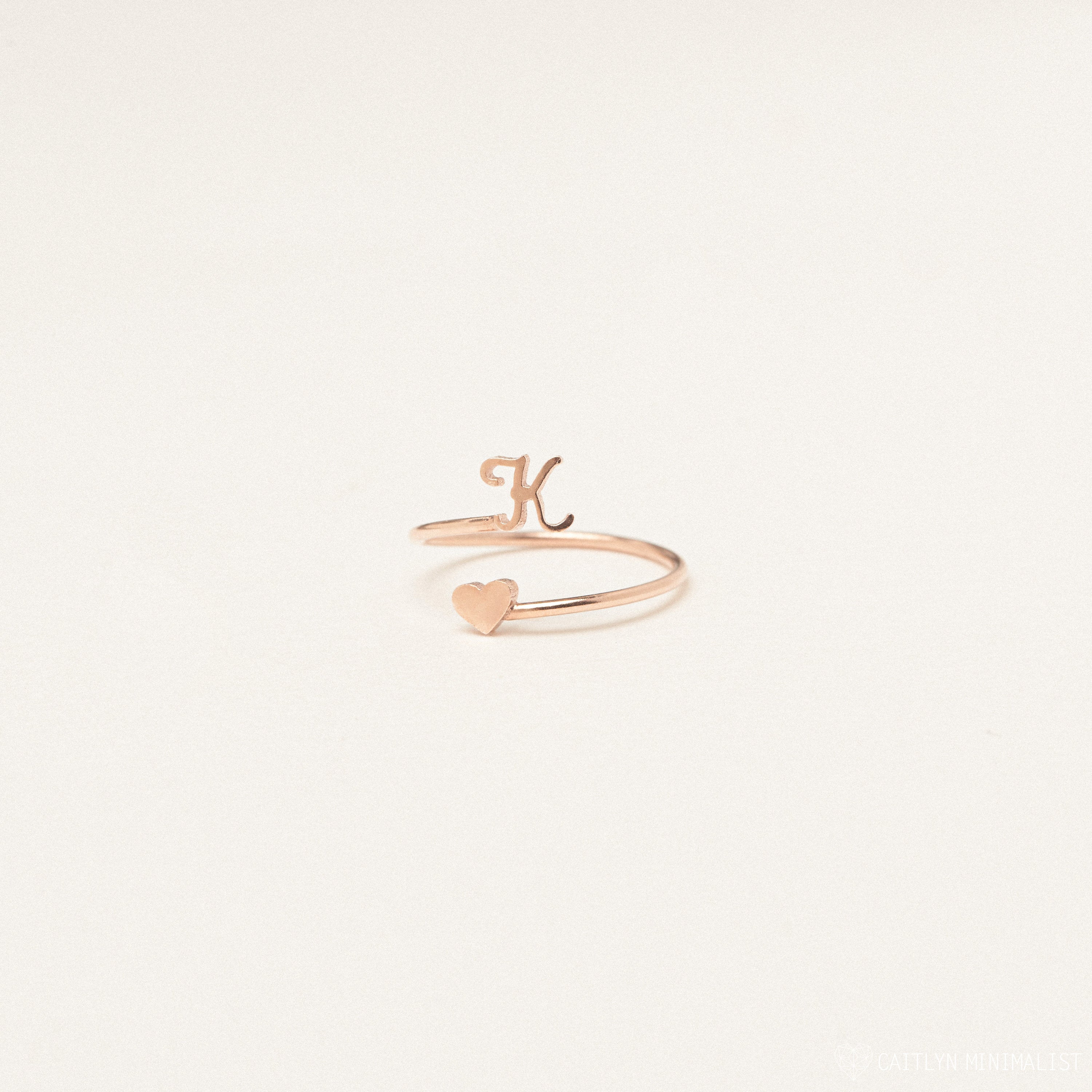 The Initial Heart Wrap Ring features a gold band with a letter K and a heart on opposite ends—an elegant symbol of love. Personalize it with any initial for a unique touch, beautifully displayed against a plain backdrop.