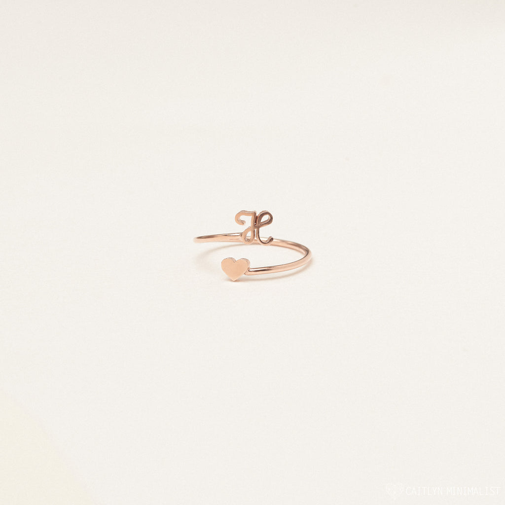 The Initial Heart Wrap Ring showcases a rose gold heart on one end and a customizable lowercase initial "e" on the other—a delicate, personalized symbol of love set against a plain background.