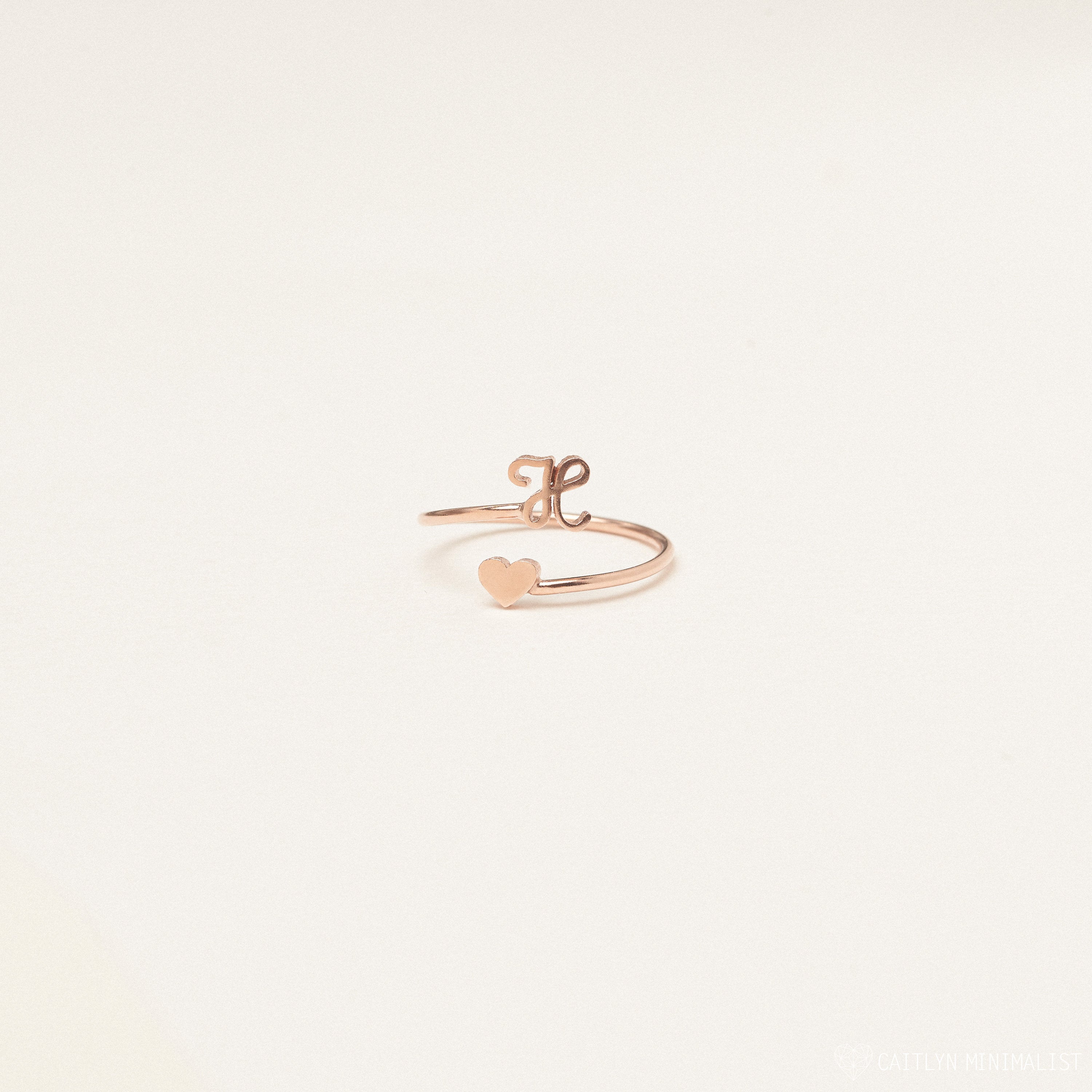 The Initial Heart Wrap Ring showcases a rose gold heart on one end and a customizable lowercase initial "e" on the other—a delicate, personalized symbol of love set against a plain background.