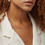 A woman wears the Zina Necklace, a double-layered gold piece, over a white textured shirt. Her curly hair frames a pearl earring.