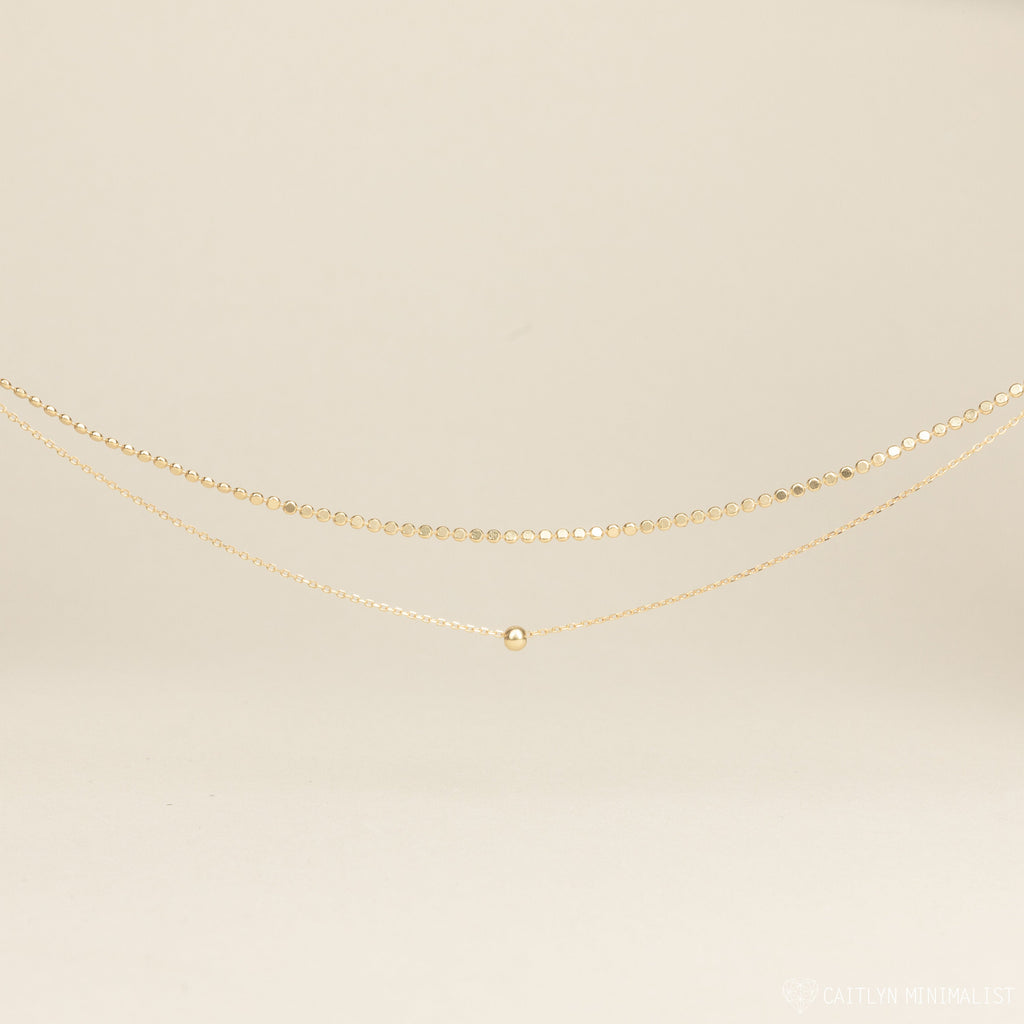 The Zina Necklace, a delicate gold chain with a single bead, is displayed on a neutral background.