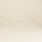 The Zina Necklace, a delicate gold chain with a single bead, is displayed on a neutral background.