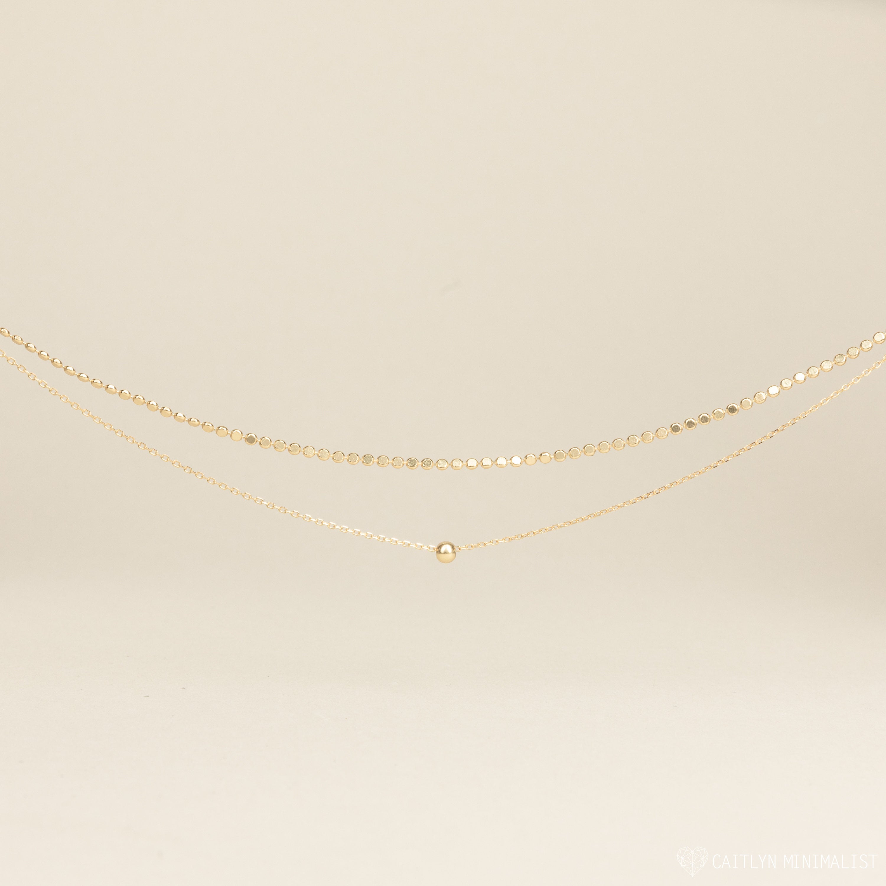 The Zina Necklace, a delicate gold chain with a single bead, is displayed on a neutral background.