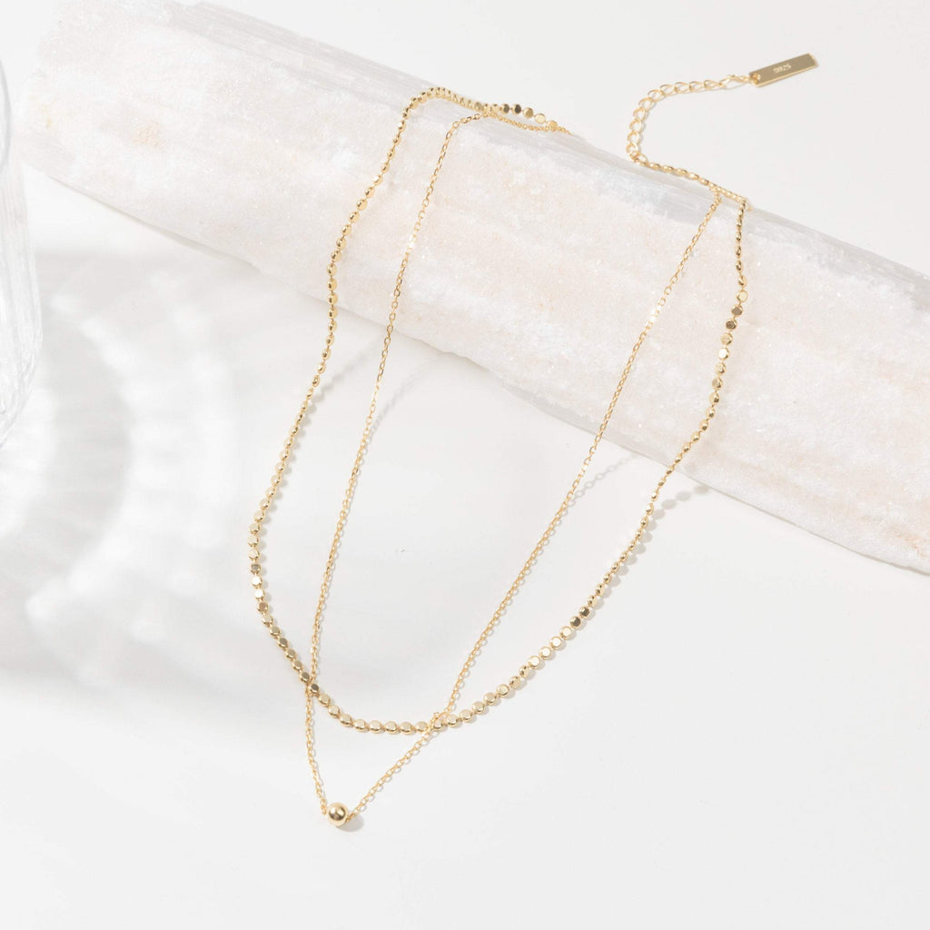 The Zina Necklace is a delicate gold double-layered piece featuring small beaded chain details, displayed on a white textured surface.
