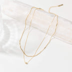 The Zina Necklace is a delicate gold double-layered piece featuring small beaded chain details, displayed on a white textured surface.