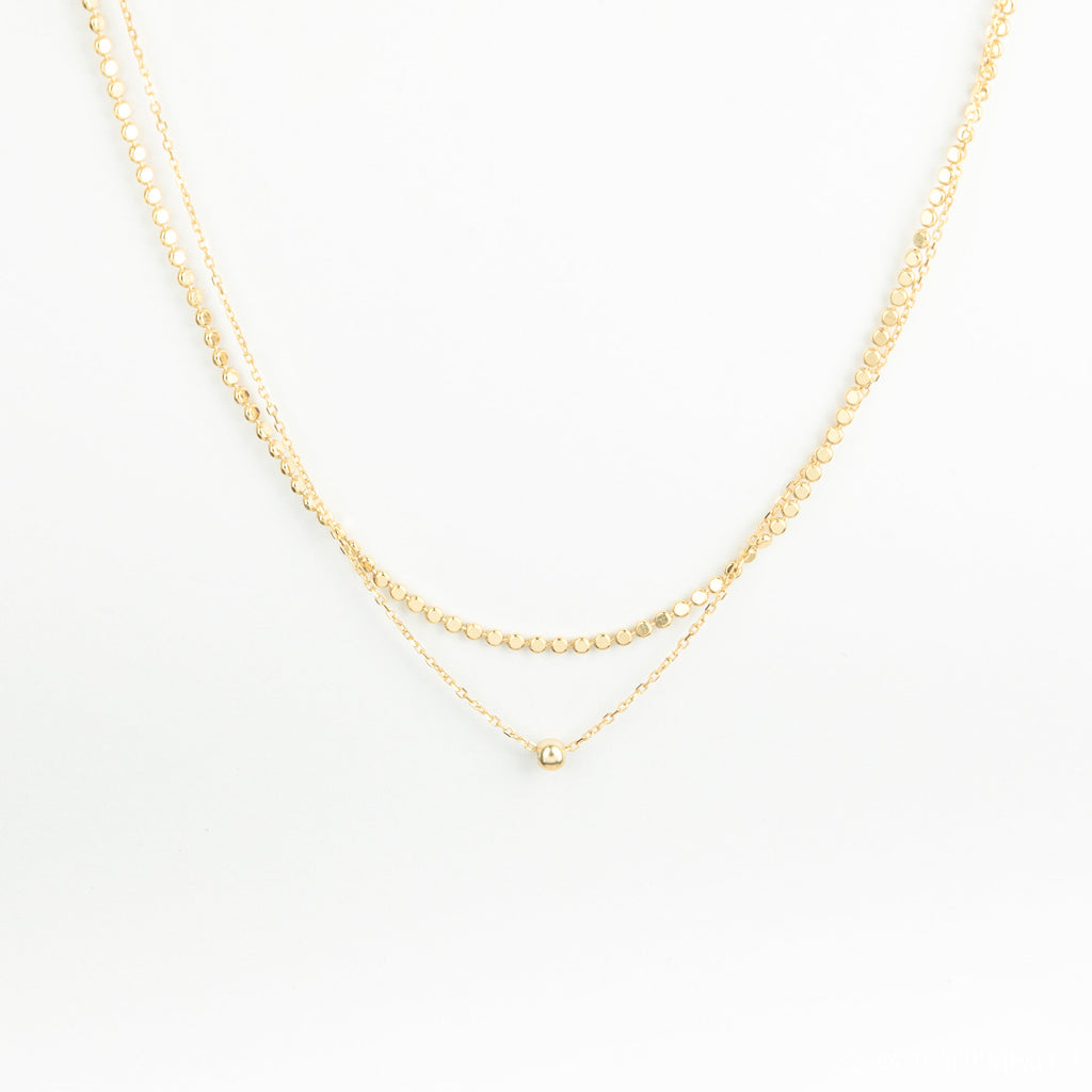 The Zina Necklace features two delicate gold chains—a double-layered design with a small gold bead and a simple pendant—displayed on a white background.