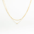The Zina Necklace features two delicate gold chains—a double-layered design with a small gold bead and a simple pendant—displayed on a white background.
