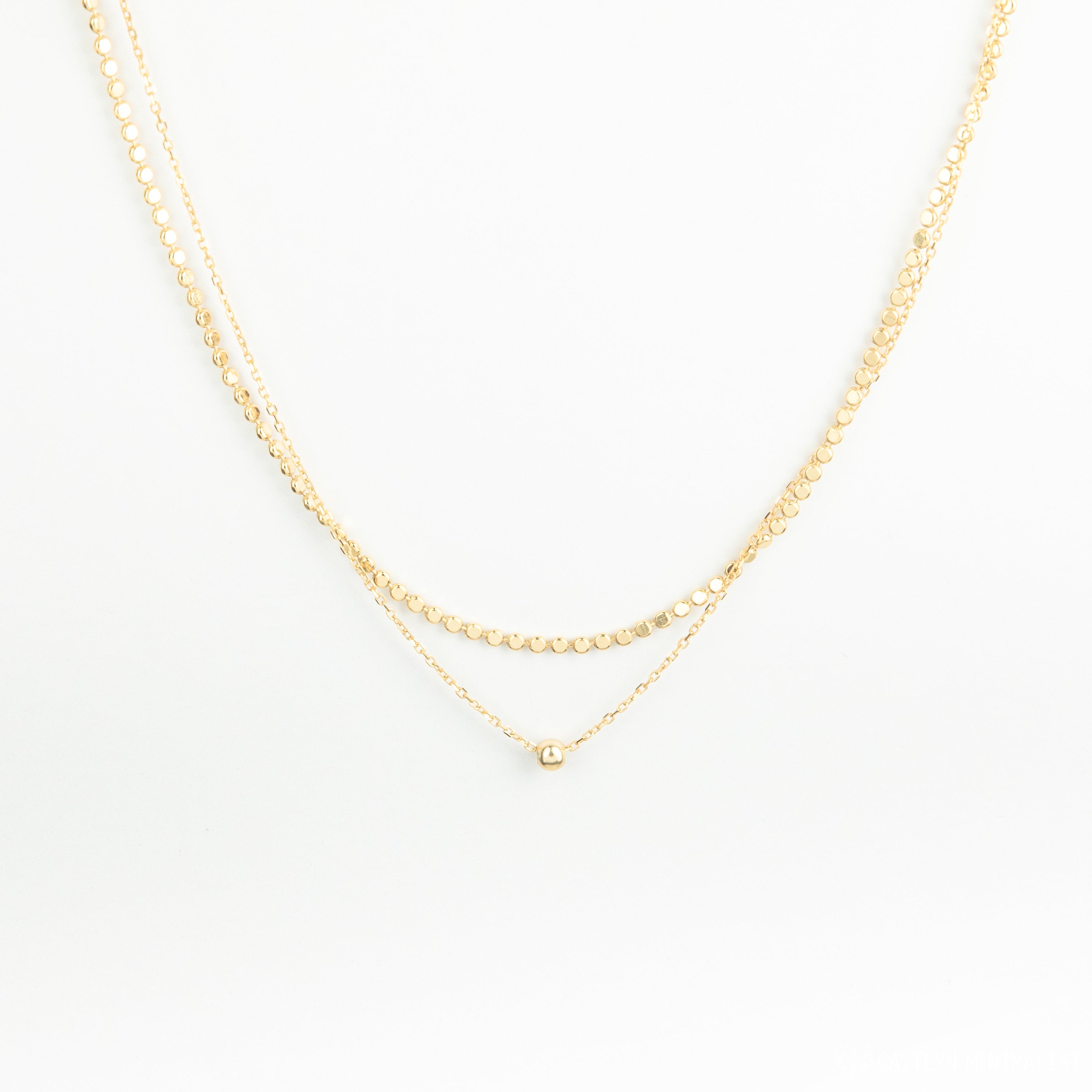 The Zina Necklace features two delicate gold chains—a double-layered design with a small gold bead and a simple pendant—displayed on a white background.