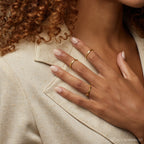 A woman with manicured nails and curly hair wears a gold Beaded Ring stacked with other bands, paired with a beige blazer.