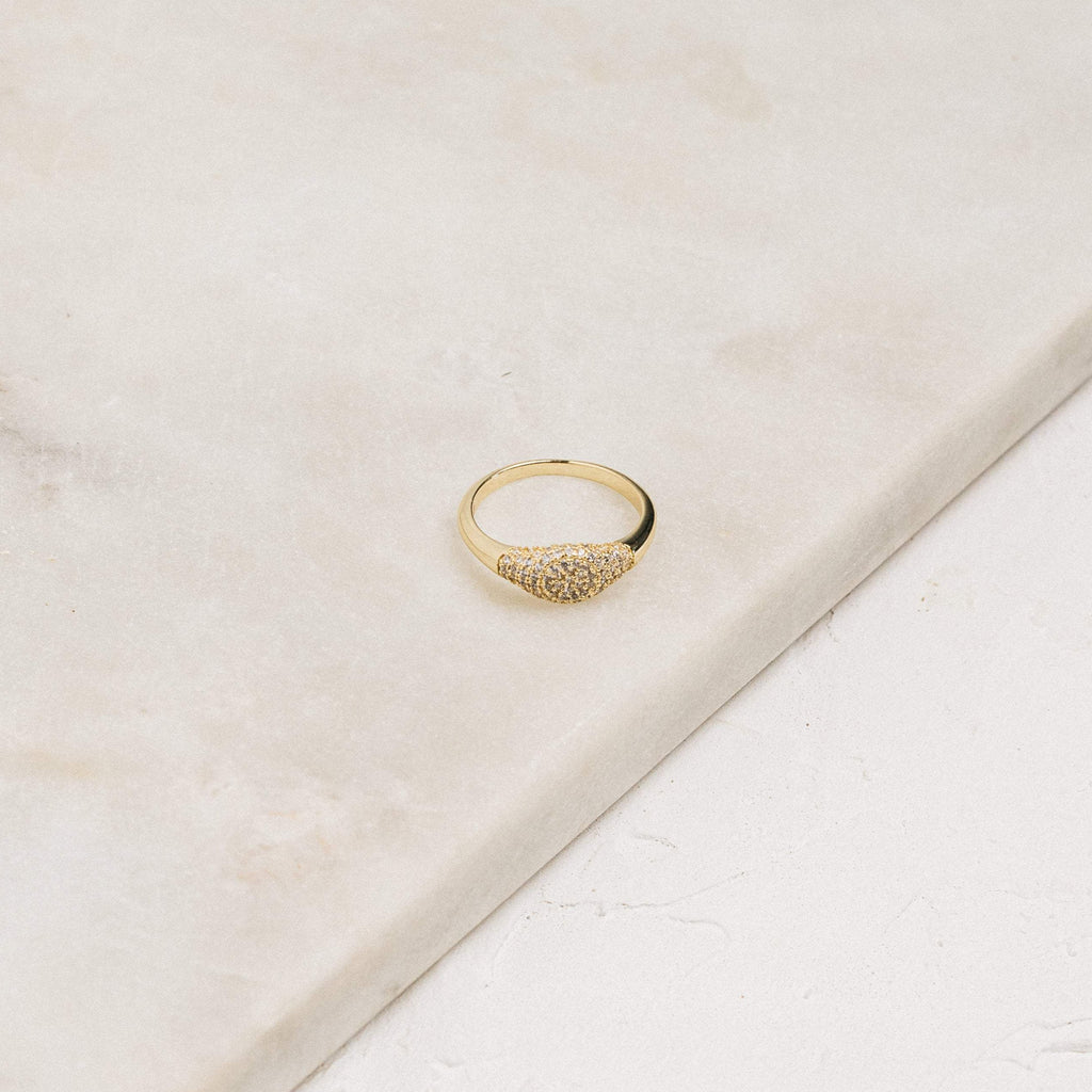 The Pave Signet Ring, adorned with a cluster of small diamonds, rests on a light marble surface, offering a modern-luxe statement accessory.