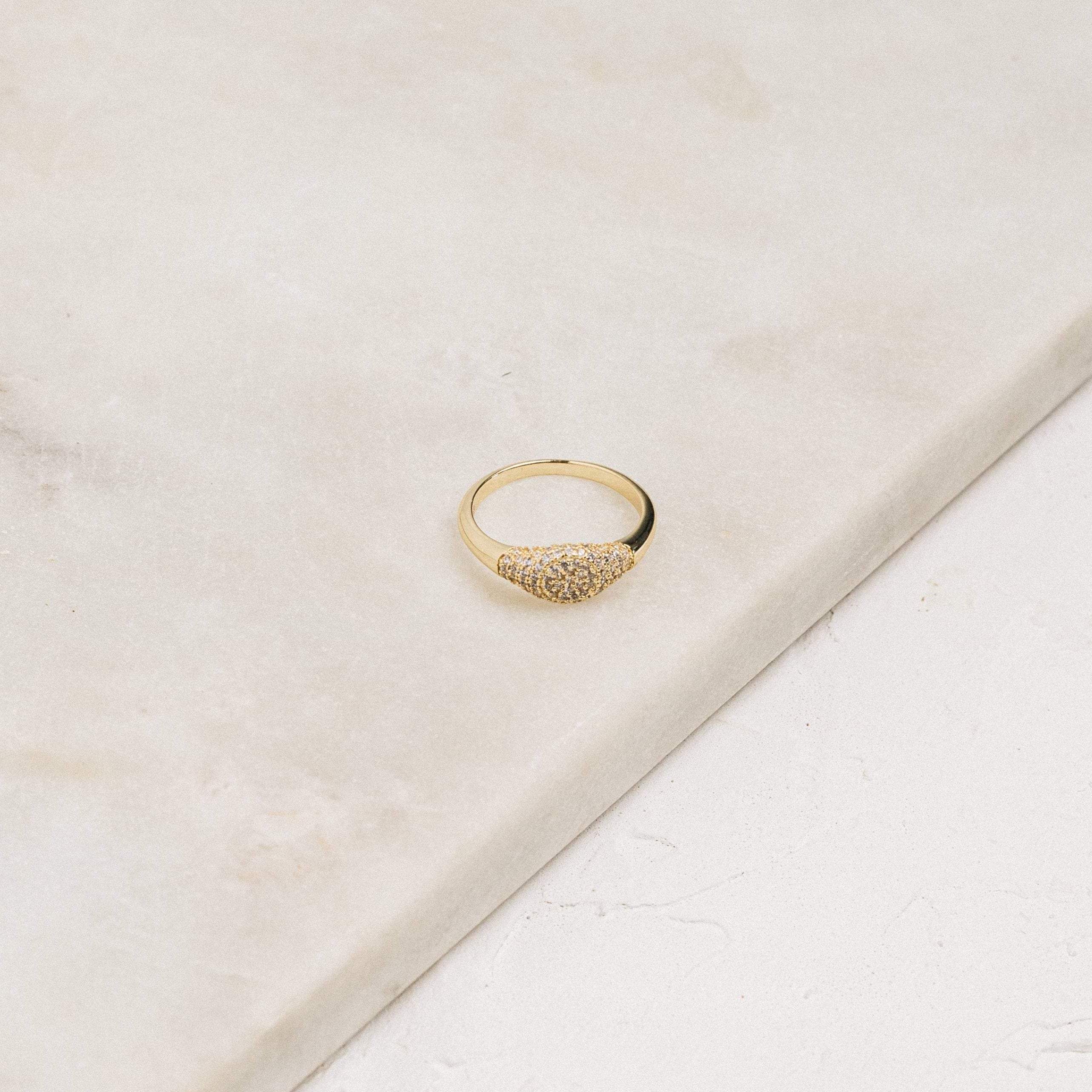 The Pave Signet Ring, adorned with a cluster of small diamonds, rests on a light marble surface, offering a modern-luxe statement accessory.