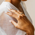 A hand wearing the Pave Signet Ring, a bold gold statement piece, rests gently on a sheer white sleeve, highlighted by neatly manicured nails for an elegant modern look.