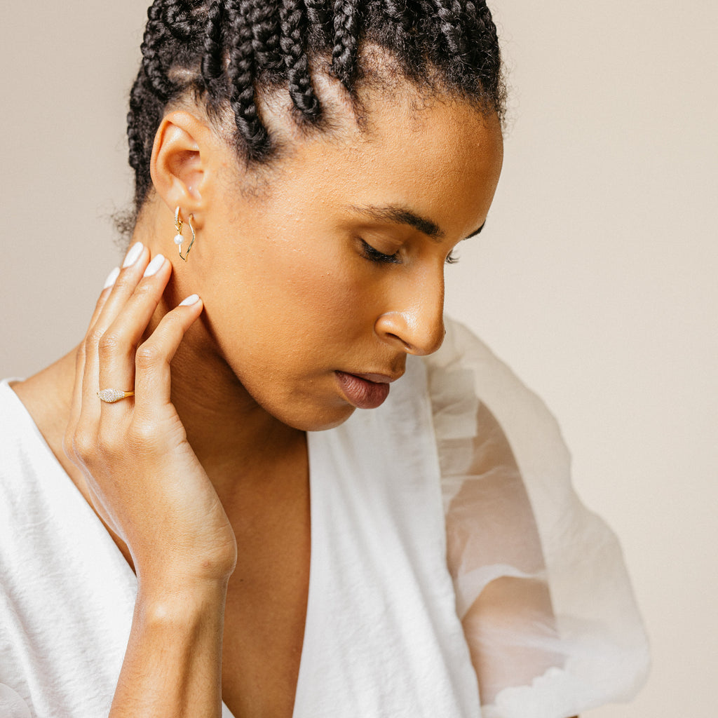 A woman with braided hair in a white top looks down, touching her Pave Signet Ring—a modern-luxe statement accessory that completes her elegant look.