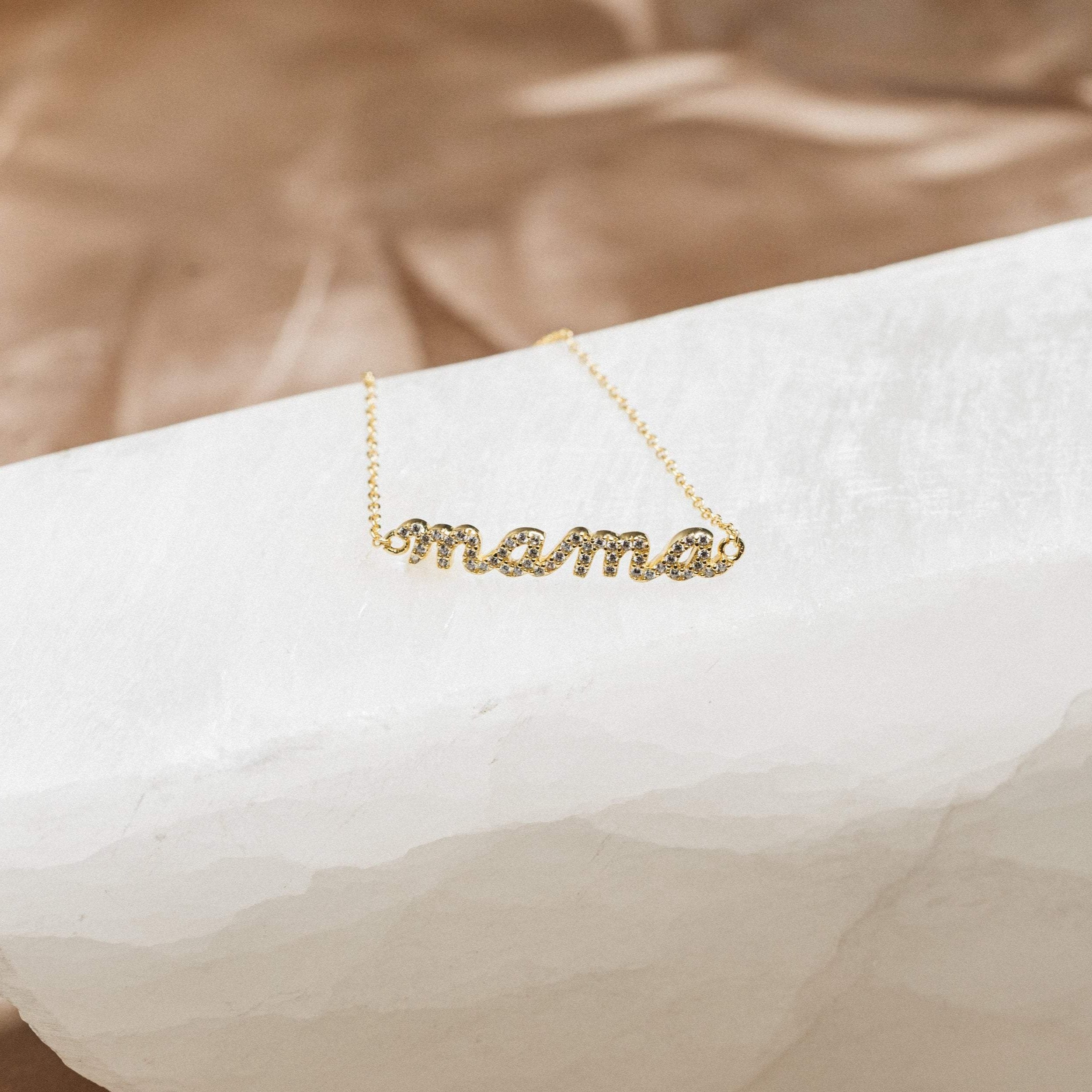 A sparkling Pave Mama Script Bracelet, featuring "mama" in elegant script and shown on a white textured surface—a timeless jewelry piece perfect for celebrating motherhood.